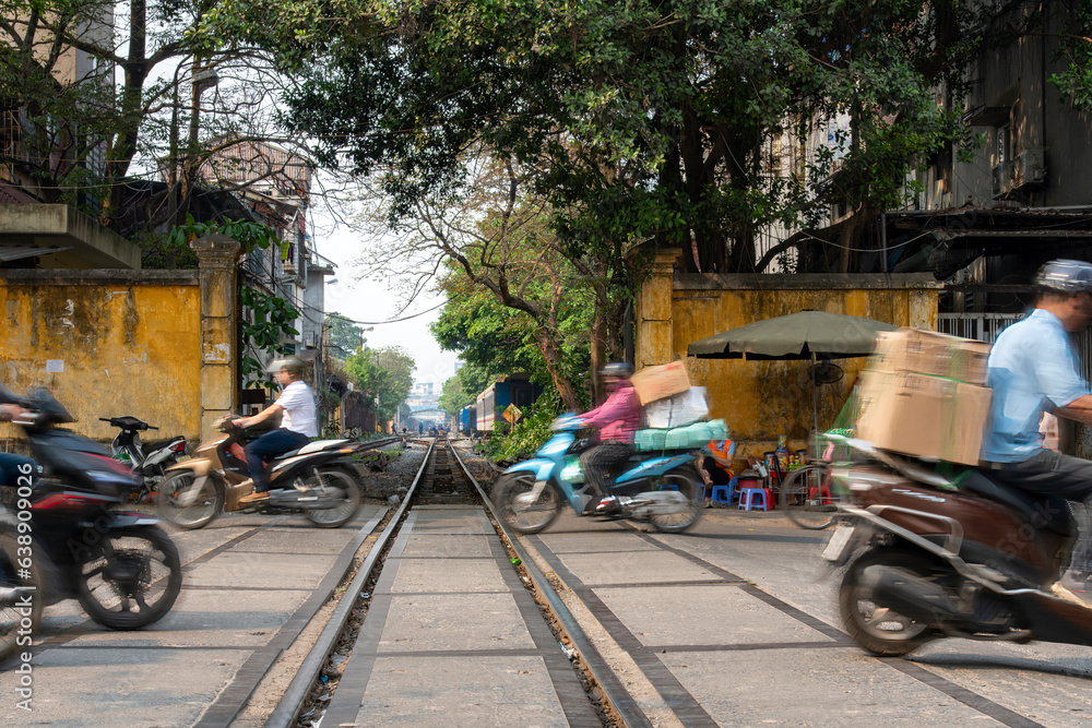 Low angle view of busy road in Hanoi, Vietnam, with scooters in blurred motion with cargo crossing the railroad tracks of the famous railroad line through center of city close to proximity of houses