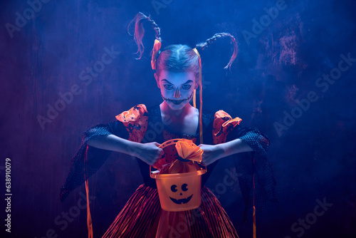 Little girl, child in costume and creepy makeup for halloween standing with basket for sweets against dark background in neon light with smoke. Concept of childhood, celebration, party, holiday, ad