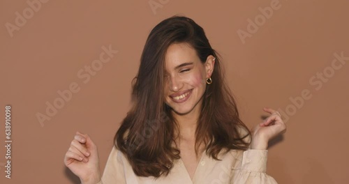 Upbeat brunette haired woman dances carefree has fun wears beige shirt, gold earrings isolated over beige background. Beautiful female model foolish around. Music, dance concept.