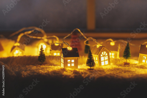 Cozy christmas miniature village. Stylish cute little glowing houses and christmas trees on soft snow blanket with lights in evening room. Atmospheric winter village still life. Merry Christmas! © sonyachny