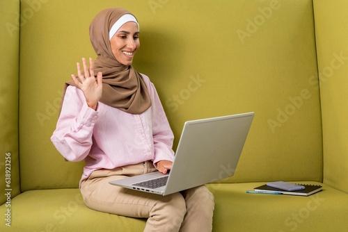 Cute Arabian Muslim woman using laptop computer, video call on sofa in coworking space, green background. Young girl watching a computer webcam waving a greeting on her hand. Remote learning or work