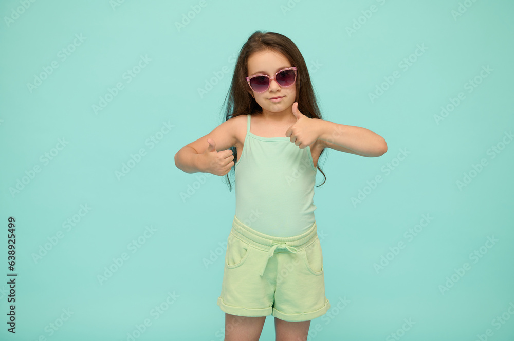 Lovely little kid girl in summer clothes and sunglasses, shows thumb up looking at camera, isolated on studio background Lovely little kid girl in summer clothes and sunglasses, shows thumb up looking at camera, isolated on studio background