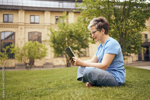 Senior woman in eyeglasses reading book while sitting on grass in park and having a rest