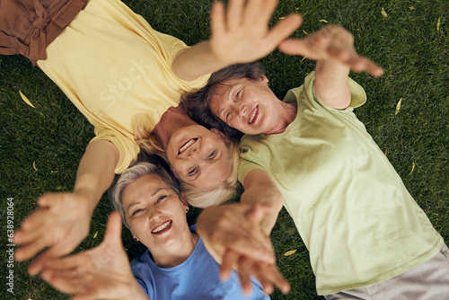 Smiling mature women friends lying head to head on grass and extend their hands to the camera