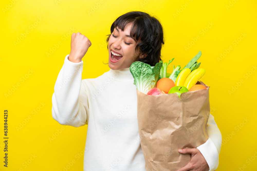 Young Argentinian woman holding a grocery shopping bag isolated on yellow background celebrating a victory Young Argentinian woman holding a grocery shopping bag isolated on yellow background celebrating a victory