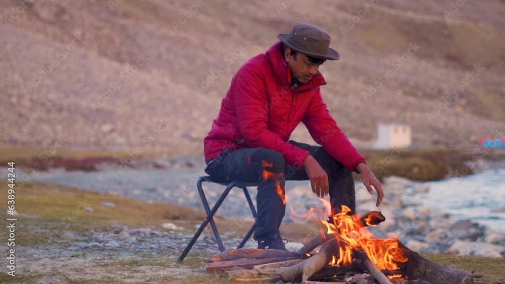 Indian male tourist fixing the woods of the bonfire in front of the mountain in Zanskar Valley at Ladakh, India. Tourist warming himself with fire in cold Himalayas in India. Indian male tourist fixing the woods of the bonfire in front of the mountain in Zanskar Valley at Ladakh, India. Tourist warming himself with fire in cold Himalayas in India.