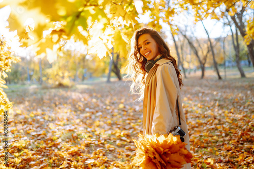 In the autumn park  a happy woman in stylish clothes and a camera is having fun among the fallen leaves. Smiling tourist woman enjoys warm sunny weather in autumn season.