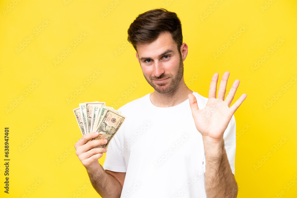 Young caucasian man taking a lot of money isolated on yellow background saluting with hand with happy expression Young caucasian man taking a lot of money isolated on yellow background saluting with hand with happy expression