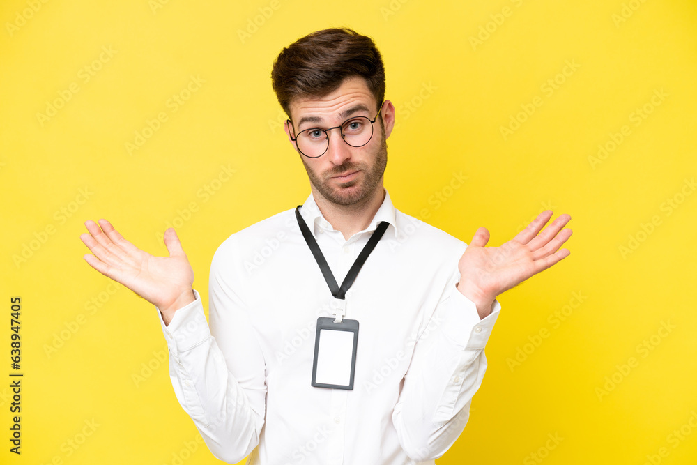 Young caucasian with ID card isolated on yellow background having doubts while raising hands Young caucasian with ID card isolated on yellow background having doubts while raising hands