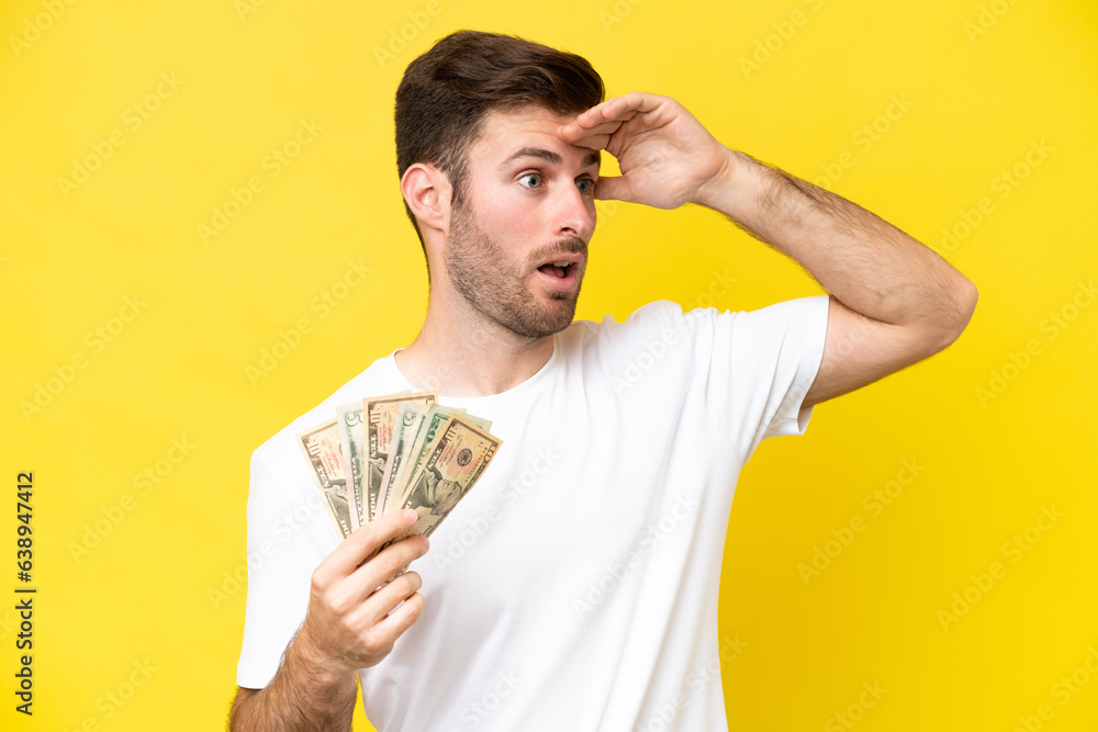 Young caucasian man taking a lot of money isolated on yellow background doing surprise gesture while looking to the side Young caucasian man taking a lot of money isolated on yellow background doing surprise gesture while looking to the side
