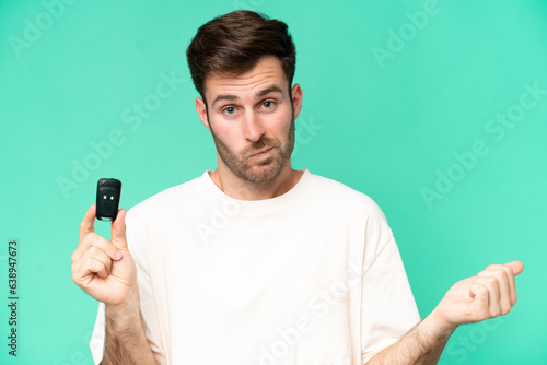 Young caucasian man holding car keys isolated on green background making doubts gesture while lifting the shoulders