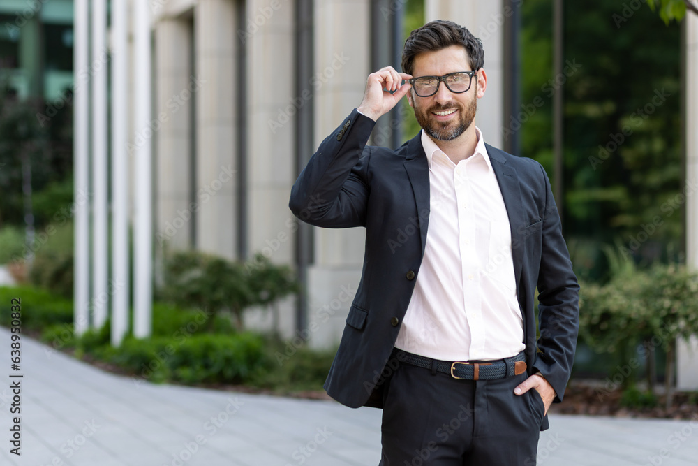 Portrait of a smiling young man in a business suit standing near an office building, holding glasses and smiling at the camera