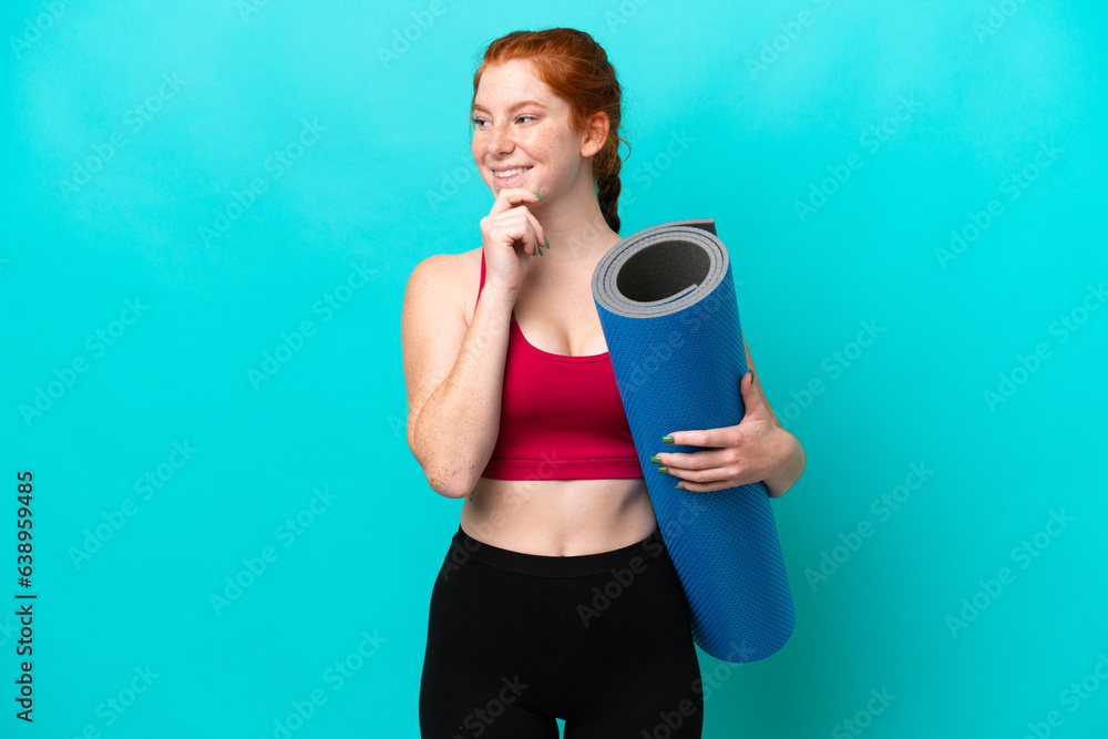 Young sport reddish woman going to yoga classes while holding a mat isolated on blue background looking to the side and smiling Young sport reddish woman going to yoga classes while holding a mat isolated on blue background looking to the side and smiling