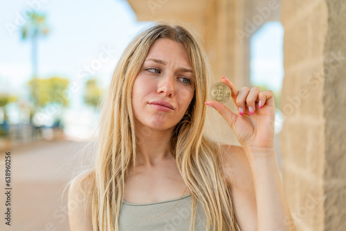 Young blonde woman holding a Bitcoin at outdoors with sad expression