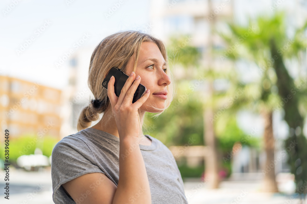 Young blonde woman at outdoors keeping a conversation with the mobile phone with someone Young blonde woman at outdoors keeping a conversation with the mobile phone with someone