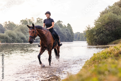Female horseback rider in a black jockey outfit riding a chestnut horse along the river at sunset. Recreation, equitation, and nature concept.