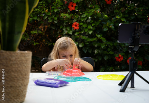 Cute blonde caucasian baby girl playing with slime and watching video lesson or cartoon on smart phone. Online education, classes at home, on distance. Child sensory development game.