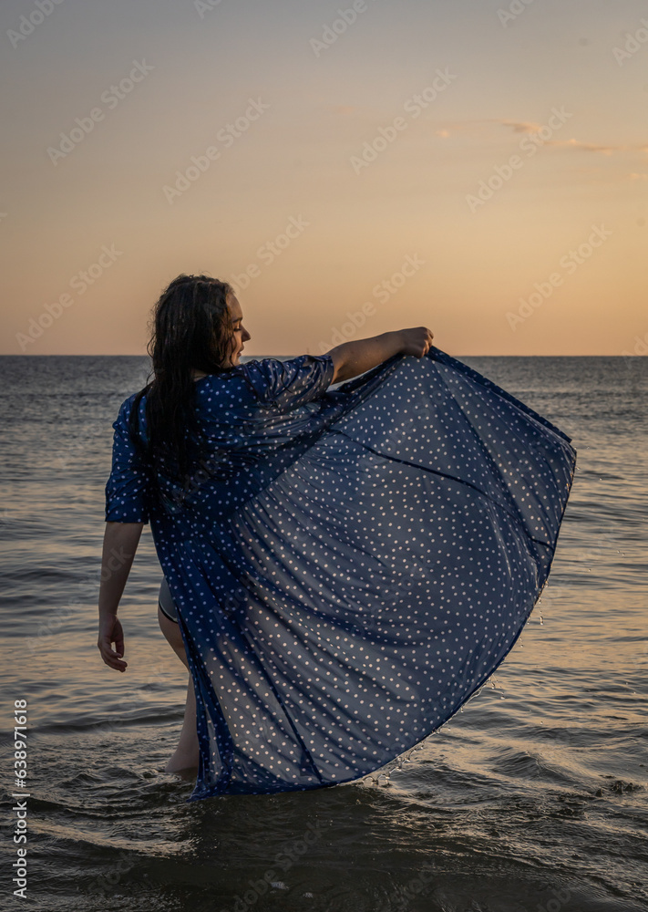 kobieta stojąca w morzu z uniesiona sukienką - obrazy, fototapety, plakaty 