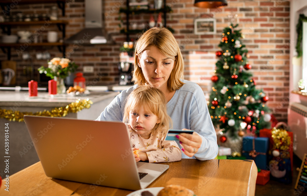 Young caucasian mother and daughter online shopping for presents for christmas and the new year holidays at home with a christmas tree behind them Young caucasian mother and daughter online shopping for presents for christmas and the new year holidays at home with a christmas tree behind them