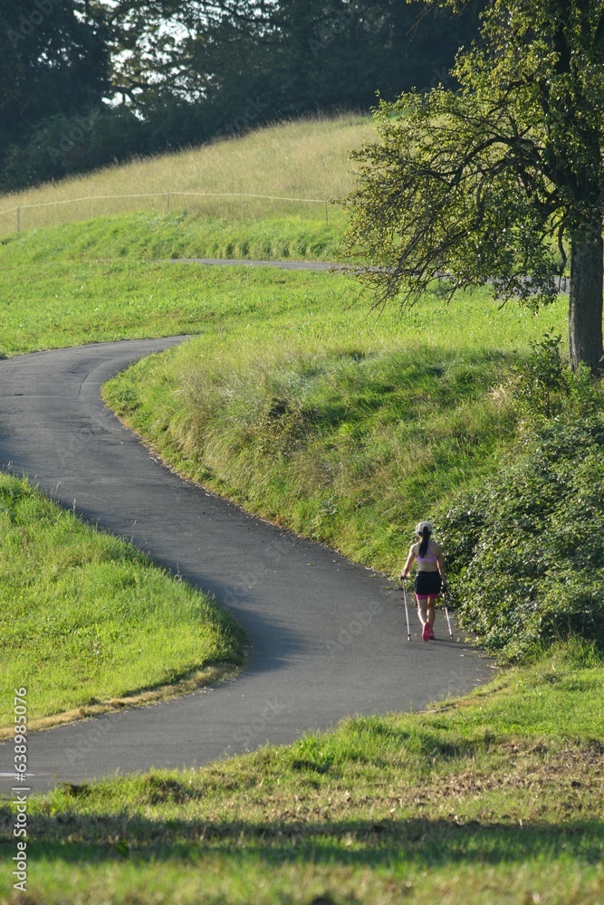 a girl walks along the road with Scandinavian sticks through the green hills