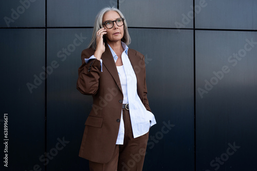 60 year old successful slender gray-haired business woman dressed stylishly speaks on the phone while walking along the wall of the business center photo