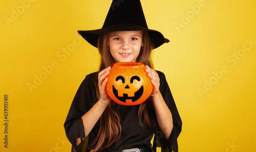 little girl with long hair smiles in a Halloween witch costume and holds a pumpkin-shaped candy bowl on a yellow background.