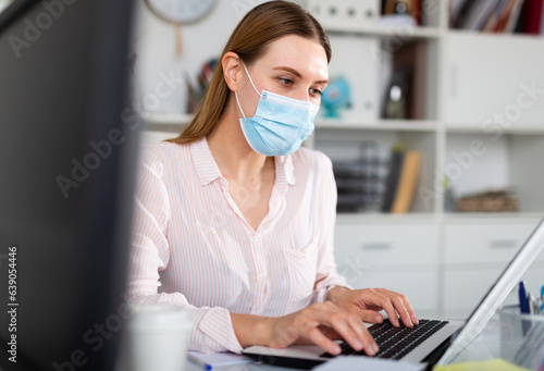 Focused young woman in disposable face mask working in business office using laptop