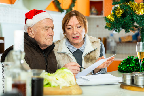 Worried mature couple sitting at table with financial documents in kitchen before christmas dinner