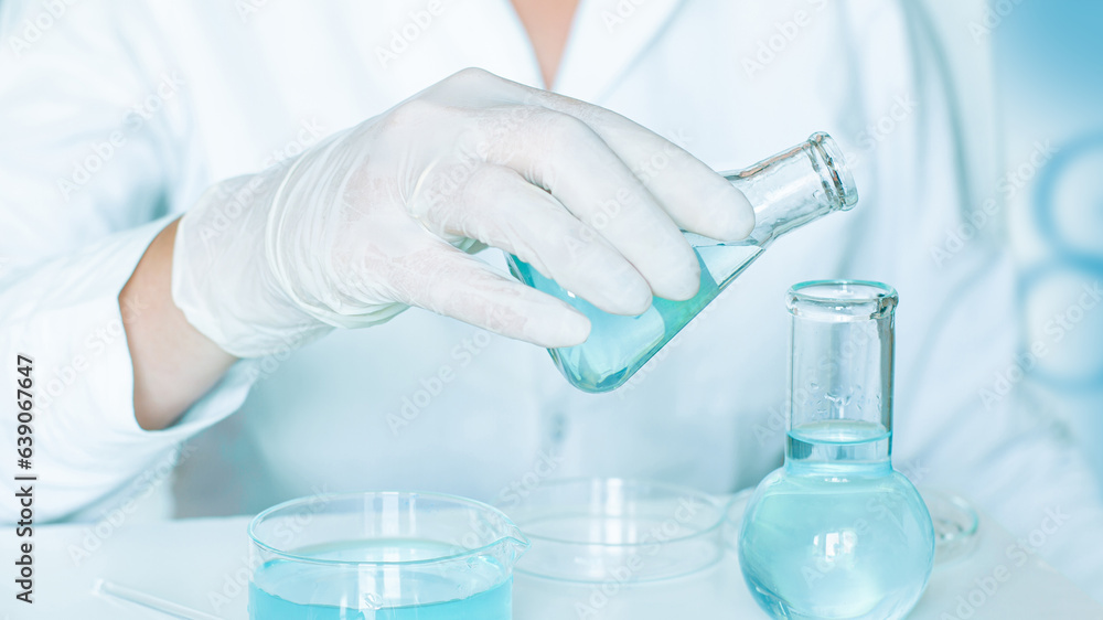 Laboratory setting: Assistant carefully fills flasks with clear blue liquid. Gloved hands. White lab coat. Glass equipment. Laboratory setting: Assistant carefully fills flasks with clear blue liquid. Gloved hands. White lab coat. Glass equipment.