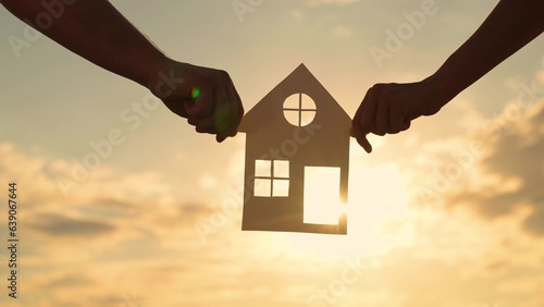 Familys hands are holding paper house at sunset, sun is shining through window. Symbol of house, happiness. Concept of building house for family. Dream to buy house. Home for children and parents, sky