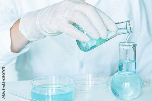 In the lab: Liquid is poured into flasks by an assistant wearing a white lab coat and gloves. Glass equipment. Liquid is clear blue.