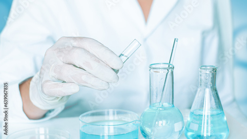 Lab environment: Aide carries out actions in the laboratory. Gloves and a white robe worn. Glassware and clear blue liquid involved.