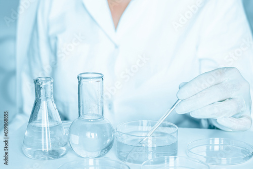 Scientific setting: Lab assistant engages in manipulations within the laboratory ambiance. Lab coat worn. Glass tools in use. Blue laboratory setting. Focused on research with transparent liquid.
