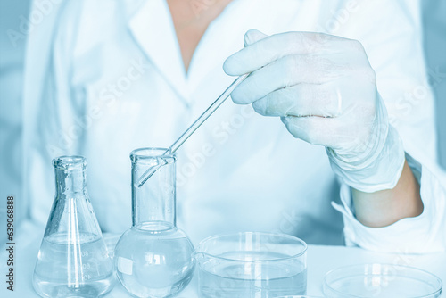 In the laboratory: Aide carefully performs actions in the laboratory surroundings. Wearing a lab coat. Utilizing glass apparatus. Situated in a blue laboratory. 