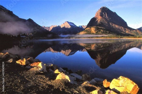 Swiftcurrent Lake - Glacier NP - MT - A mirror-like blue lake reflects the buttes on the far shore. - On the left shore of the lake a lodge is veiled by fog.