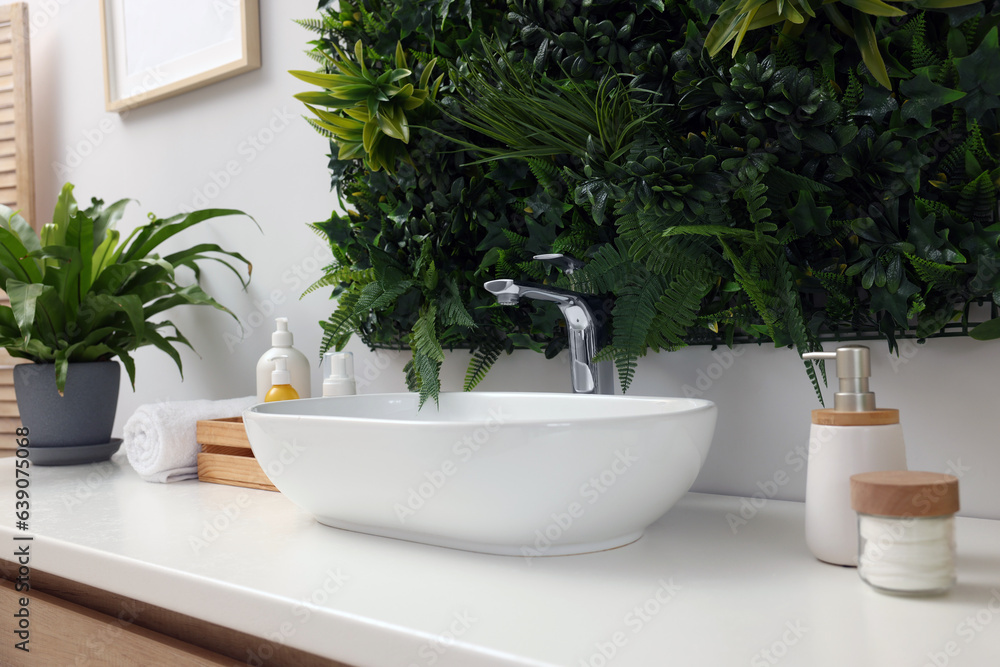 Green artificial plants, vessel sink and different personal care products in bathroom Green artificial plants, vessel sink and different personal care products in bathroom