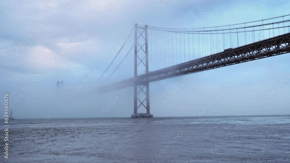View of 25 de Abril Bridge famous tourist landmark of Lisbon connecting Lisboa and Almada in heavy fog mist wtih yacht boats passing under. Lisbon, Portugal View of 25 de Abril Bridge famous tourist landmark of Lisbon connecting Lisboa and Almada in heavy fog mist wtih yacht boats passing under. Lisbon, Portugal