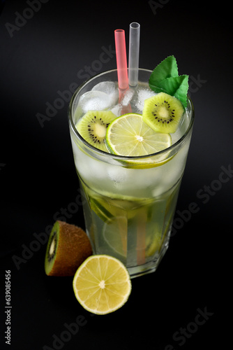 A tall glass of lime juice with kiwi, with ice, tubules and mint leaves on a black background, next to the ripe fruit.