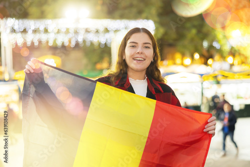 Smiling young traveler girl in warm red and black plaid shirt enjoying walk on traditional street Christmas fair in European town, waving national flag of Belgium ..