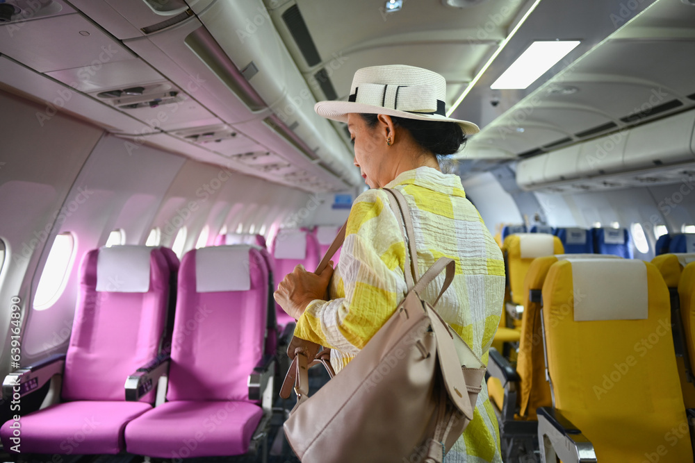 Smiling middle age female tourist with bag walking the aisle on plane. Retirement, travel and summer vacation concept Smiling middle age female tourist with bag walking the aisle on plane. Retirement, travel and summer vacation concept