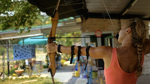 Sport woman in shooting range with bow. Sportsman in shooting gallery aim an arrow to hit target. Outdoor archery training. Practice and training of archery in shooting range. Athlete keep wooden bow