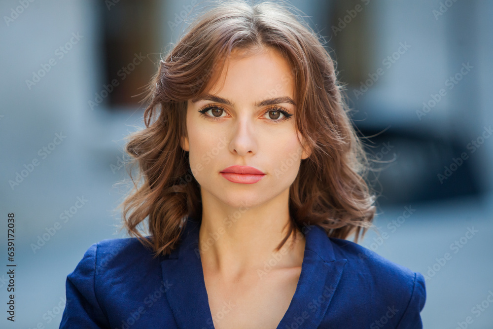 Portrait of a beautiful young woman with long brown hair, dressed in a blue jacket, standing against the background of an office building. strong and confident business woman Portrait of a beautiful young woman with long brown hair, dressed in a blue jacket, standing against the background of an office building. strong and confident business woman