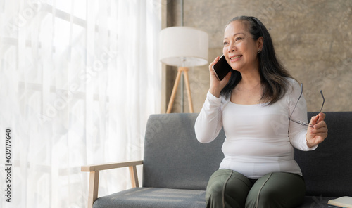 Happy Asian senior woman holding mobile phone for online video call Sitting on the sofa, call family members using mobile applications.