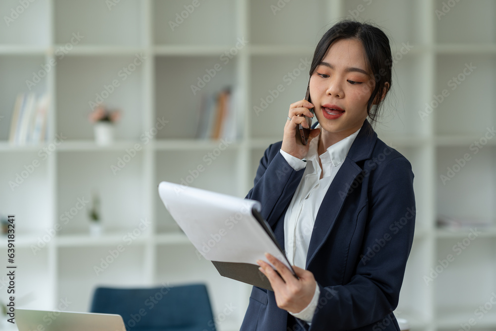 Asian businesswoman standing working with laptop computer, mobile phone, tablet to contact, chat, search internet, news, email, chat while working on finance, marketing, accounting data in office. Asian businesswoman standing working with laptop computer, mobile phone, tablet to contact, chat, search internet, news, email, chat while working on finance, marketing, accounting data in office.