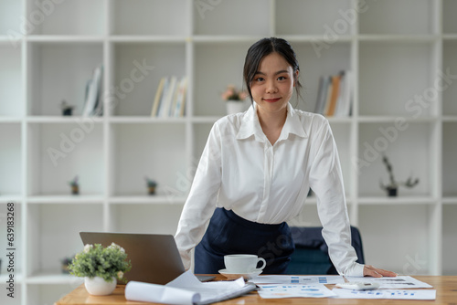 Asian businesswoman standing working with laptop computer, mobile phone, tablet to contact, chat, search internet, news, email, chat while working on finance, marketing, accounting data in office.