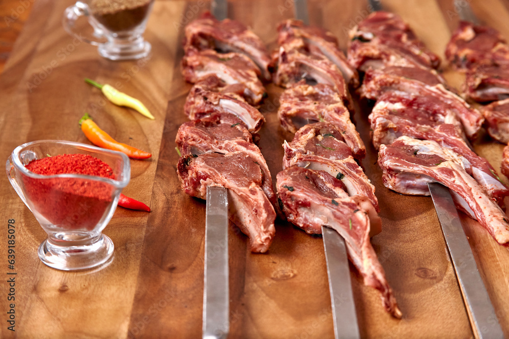 Metal skewers with raw uncooked lamb ribs meat for frying on the wooden board with chili peppers, garlic and spices near it. Close-up perspective view, shallow depth of field. Meat in focus