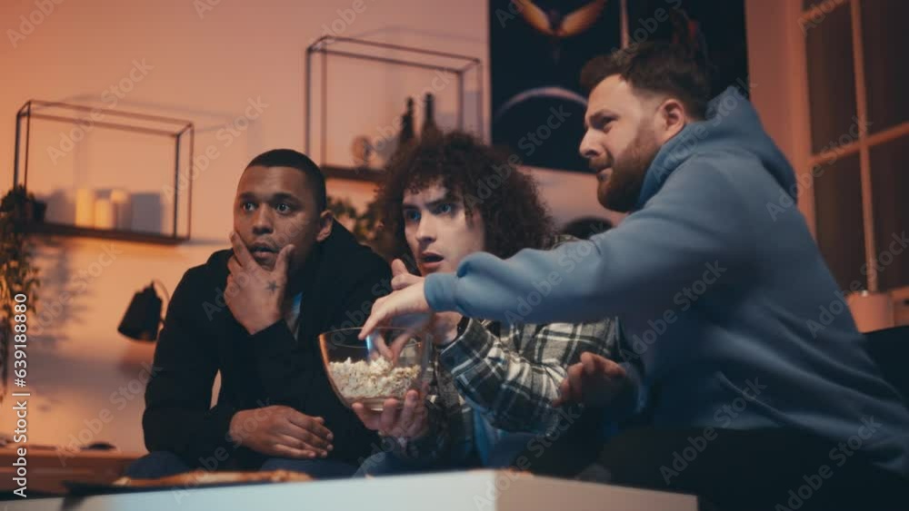 Three male friends watching a scary horror movie at home, eating popcorn Three male friends watching a scary horror movie at home, eating popcorn