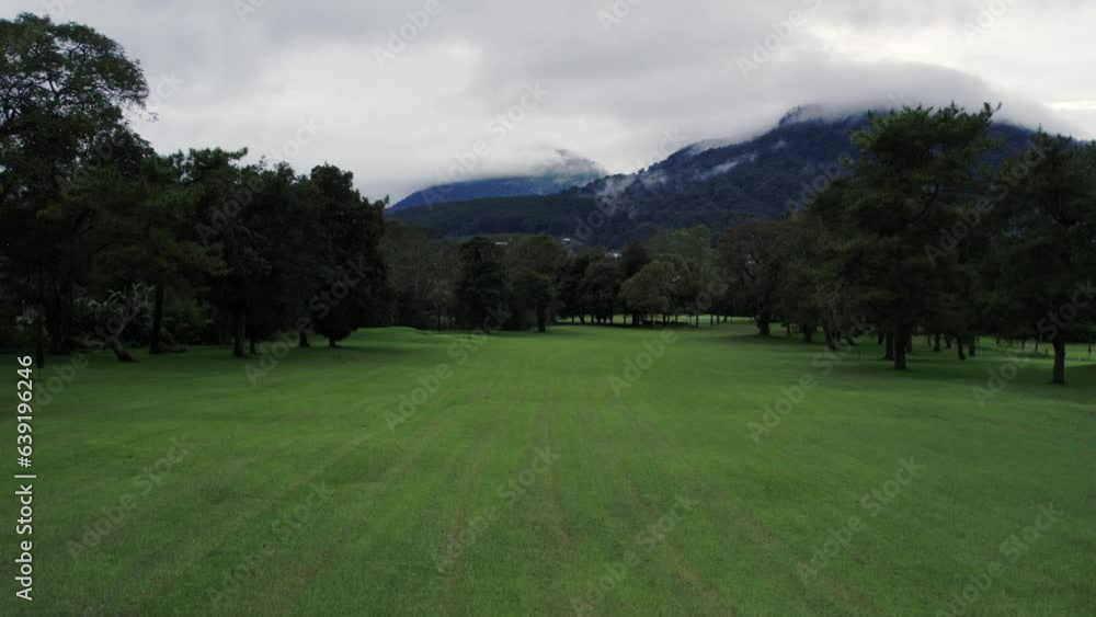 Aerial view of golf course and village with houses near lake, green trees and field. Drone flying down a recreational place, mountain and cloudy sky. Concept of holiday and island Aerial view of golf course and village with houses near lake, green trees and field. Drone flying down a recreational place, mountain and cloudy sky. Concept of holiday and island