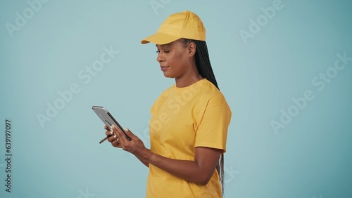 Delivery woman in uniform holding tablet, writing something and looking at the screen focused. Isolated on blue background. photo