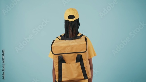 Delivery woman in uniform with portable backpack refrigerator, posing at the camera with her back. Isolated on blue background. photo